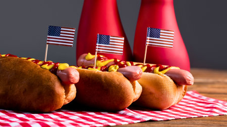 Three delicious hot dogs adorned with American flags sit on a red and white checkered cloth, with ketchup bottles in the background.の写真素材