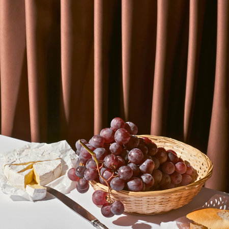 A still life arrangement featuring a basket of red grapes, cheese wedges, a knife, and a textured curtain in the background.の写真素材