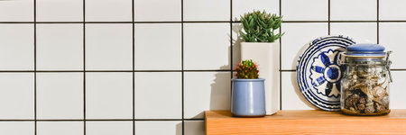 A minimalist arrangement of two small potted plants, a decorative plate, and a jar on a wooden shelf. The background is a white tiled wall.の写真素材