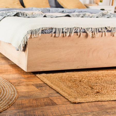 A close-up view of a wooden bed frame with a fringed blanket and a woven rug on a wooden floor.の写真素材