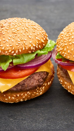Close-up view of two gourmet burgers with sesame seed buns, cheese, lettuce, tomato, and red onion on a dark surface.の写真素材