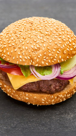 A close-up shot of a delicious cheeseburger featuring a beef patty, melted cheese, red onion, lettuce, and tomato on a sesame seed bun.の写真素材