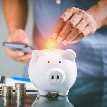 A person is placing coins into a white piggy bank. A smartphone and stacks of coins are also visible.の写真素材