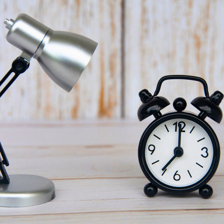 A silver desk lamp shines light on a classic black twin-bell alarm clock. The background is a light-colored wooden texture.の写真素材