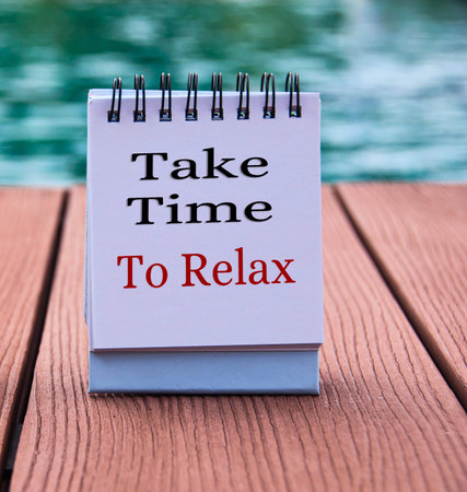 A small notepad with the words Take Time To Relax sits on a wooden surface near a swimming pool, encouraging relaxation.の写真素材