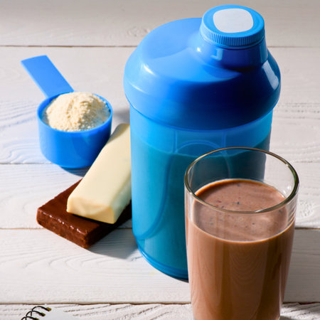 A blue shaker bottle, scoop of protein powder, chocolate bar, and a glass of chocolate protein shake on a wooden surface.の写真素材