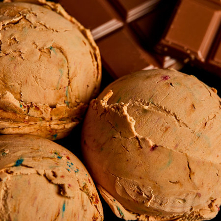 A close-up shot shows three generous scoops of light brown ice cream, possibly caramel or coffee flavored, with hints of colorful sprinkles. Chocolate bars are visible in the background.の写真素材