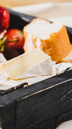 A close-up shot of creamy Brie cheese and a slice of rustic bread, artfully arranged on a dark wooden board with a hint of red fruit.の写真素材