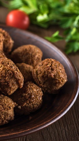 Close-up of golden brown falafel balls arranged on a dark, textured plate, with blurred green herbs and a red tomato in the background.の写真素材