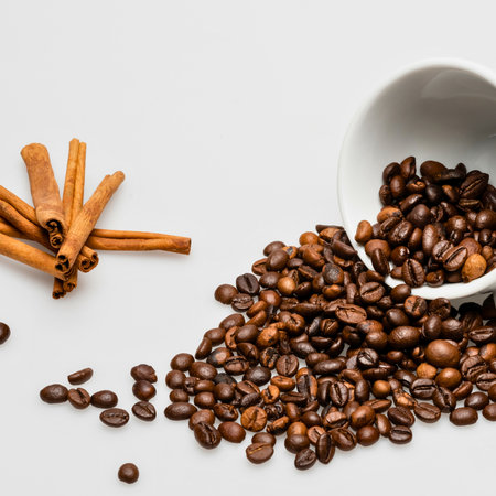 A white bowl spills roasted coffee beans and scattered beans next to a pile of cinnamon sticks on a plain white surface.の写真素材