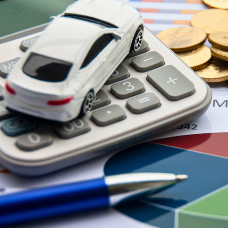 A white toy car sits on a calculator surrounded by coins and financial charts, symbolizing car financing and budgeting.の写真素材