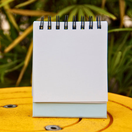 A small, blank spiral-bound notebook sits open on a textured yellow surface. Lush green foliage is visible in the background.の写真素材