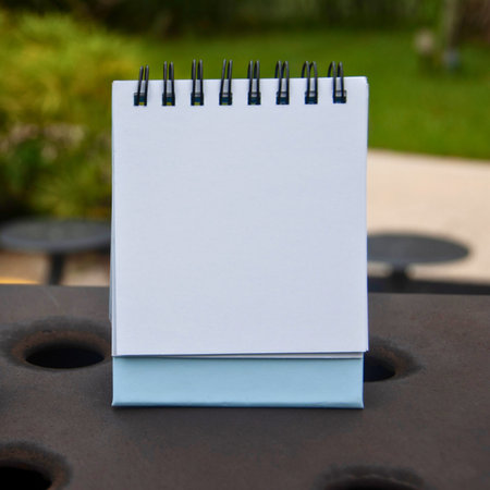 A small, white, blank spiral notebook stands open on a dark, textured surface with a blurred green background.の写真素材
