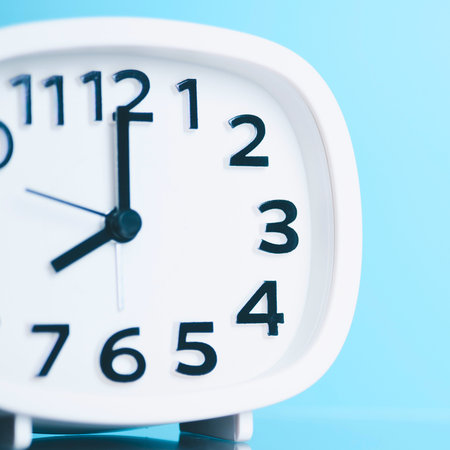 A white analog alarm clock with black numbers and hands is set to 8:00 against a soft blue background.の写真素材
