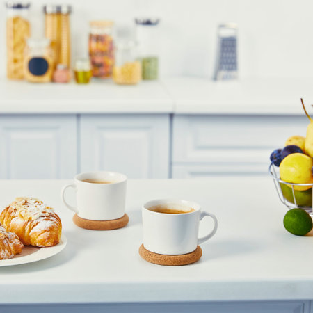 Two mugs of coffee and a croissant sit on a white counter, with a fruit basket in the background.の写真素材