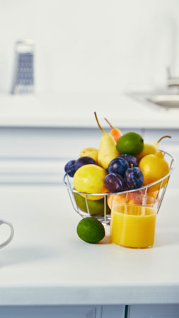 A vibrant assortment of fresh fruits fills a glass bowl next to a yellow citrus juicer on a clean kitchen counter.の写真素材