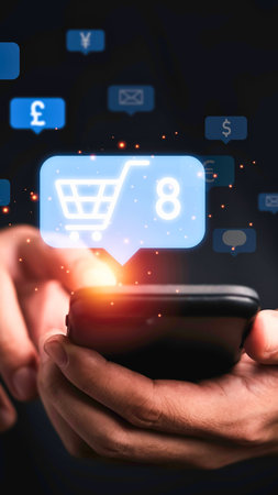 Hands holding a smartphone displaying a glowing shopping cart icon with a number eight and various currency symbols in the background.の写真素材