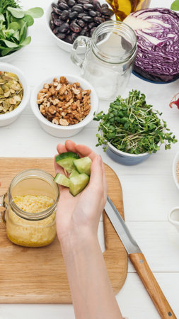 A hand is adding avocado to a jar of dressing, surrounded by various healthy ingredients like greens, nuts, and seeds.の写真素材