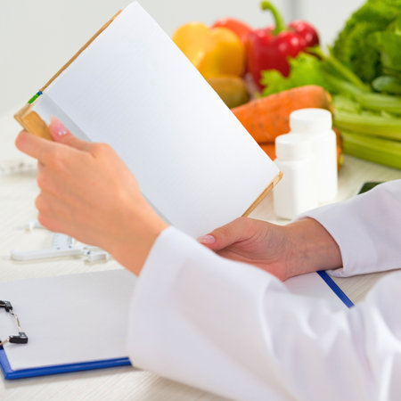 A doctor or nutritionist in a white coat holds a document, with a vibrant assortment of fresh vegetables and a supplement bottle visible.の写真素材