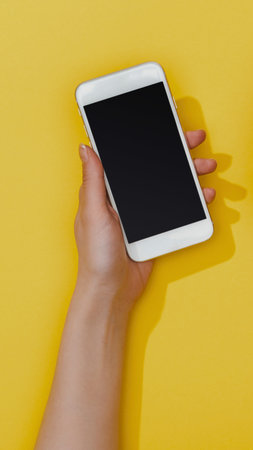 A person's hand holds a white smartphone with a blank black screen against a vibrant yellow surface, casting a soft shadow.の写真素材