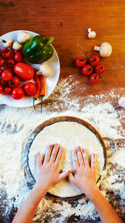 A top-down view shows hands pressing pizza dough on a wooden table dusted with flour. Fresh tomatoes and basil are visible.の写真素材