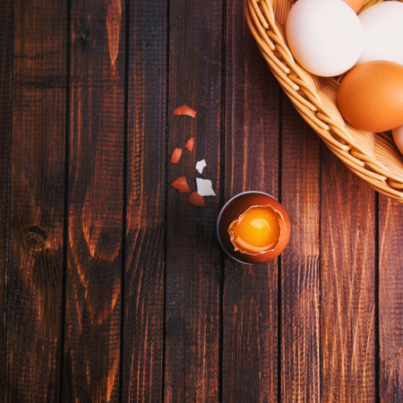 A basket of fresh eggs rests on a dark wooden surface next to a cracked egg with its yolk exposed.の写真素材