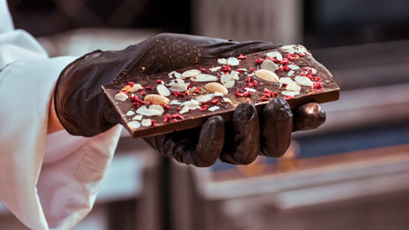 A gloved hand holds a freshly made, dark chocolate bar topped with a colorful mix of nuts and dried berries.の写真素材