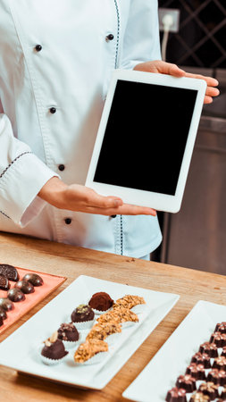 A chef in a white uniform presents a tablet computer above a selection of beautifully arranged artisanal chocolates.の写真素材