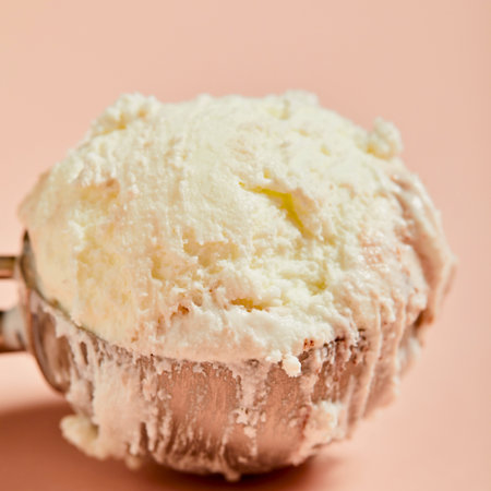 A close-up shot of a perfectly scooped ball of creamy vanilla ice cream melting slightly on a metal scoop against a soft pink background.の写真素材