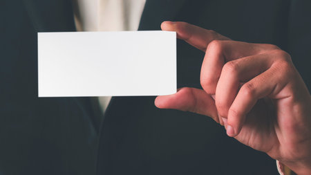 A close-up shot of a person's hand holding a blank white business card. The background is a dark suit.の写真素材