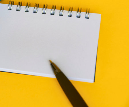 A close-up shot of a blank spiral-bound notebook page with a pen poised above it, set against a bright yellow surface.の写真素材