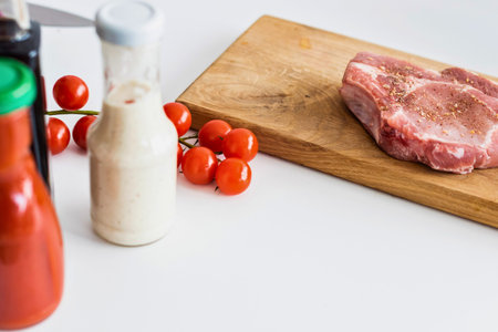 A raw pork chop sits on a wooden cutting board next to cherry tomatoes and condiment bottles on a...