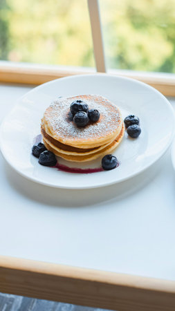 A close-up shot of golden brown pancakes topped with fresh blueberries and a drizzle of syrup, presented on a white plate.の写真素材