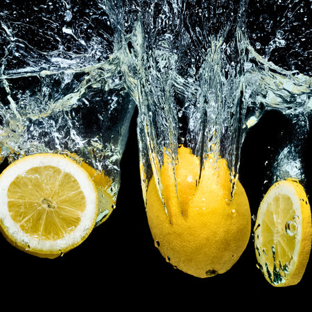 Three bright yellow lemons, one sliced and two whole, plunge into dark water creating a dramatic splash and many water droplets.の写真素材