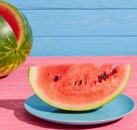 A vibrant slice of juicy watermelon rests on a blue plate, with a whole watermelon in the background, set against a pink and blue wooden surface.の写真素材