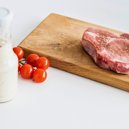 A raw pork chop sits on a wooden cutting board next to cherry tomatoes and a glass of milk, ready for cooking.の写真素材