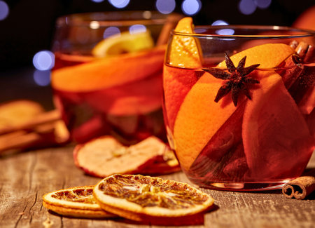 Close up of festive mulled wine in glasses, garnished with orange slices, apples, and star anise, with bokeh lights in the background.の写真素材