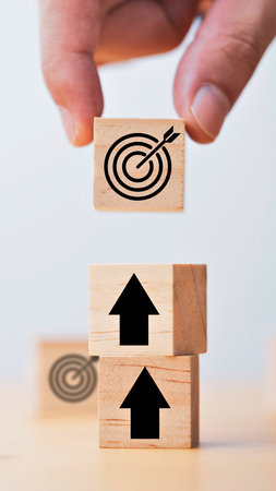 A hand carefully places a wooden block with a target symbol on top of two blocks featuring upward arrows, representing reaching goals.の写真素材
