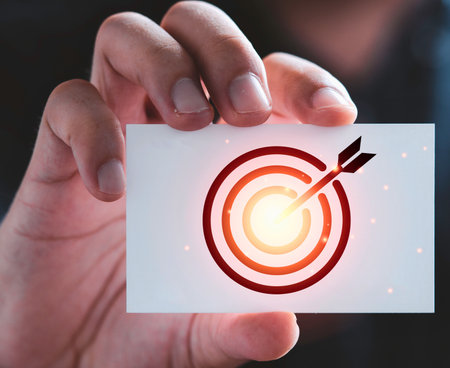 A close-up shot shows a hand holding a white business card featuring a red target with an arrow hitting the bullseye.の写真素材