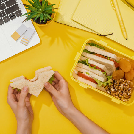 Hands assemble a sandwich for a packed lunch, surrounded by office supplies and a small green plant on a vibrant yellow surface.の写真素材