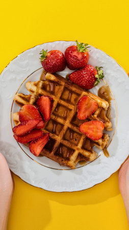 A golden brown waffle is generously topped with sliced fresh strawberries, presented on a decorative white plate against a vibrant yellow background.の写真素材