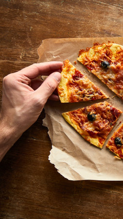 A hand picks up a slice of pizza from a rustic wooden table. The pizza has olives and red sauce.の写真素材