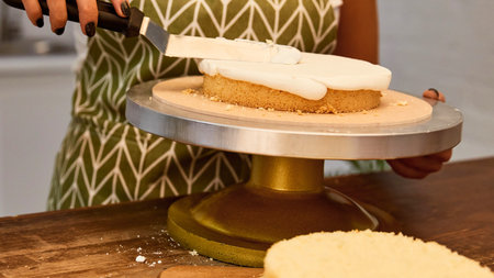 A person in an apron is carefully frosting a cake on a rotating stand, smoothing the icing with a spatula.の写真素材