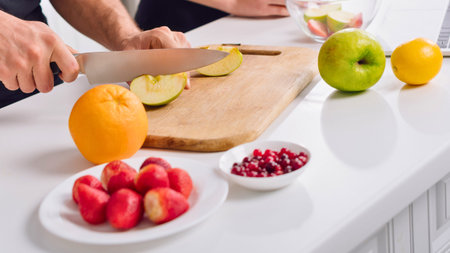 Hands slicing apples on a cutting board with an assortment of fresh fruits including oranges, rose apples, and pomegranate seeds.の写真素材
