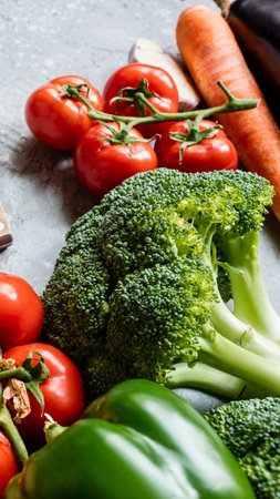 A vibrant collection of ripe tomatoes, broccoli, green bell pepper, carrots, and eggplant arranged on a light gray, textured background.の写真素材