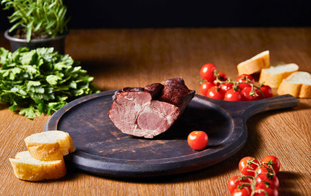 A dark slate serving board displays cured meat, cherry tomatoes, and bread on a wooden table, with fresh herbs in the background.の写真素材