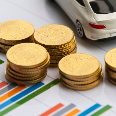 Stacks of golden coins are placed on financial charts next to a white car, symbolizing wealth and automotive investment.の写真素材