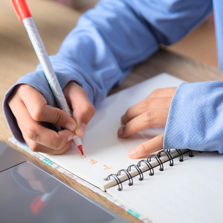 A person's hands are shown writing in a spiral-bound notebook with a red and white pen, focused on...