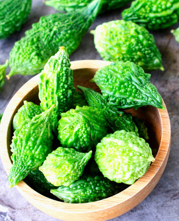 A close-up shot of vibrant green bitter melons, some piled in a rustic wooden bowl and others scattered on a textured surface.の写真素材