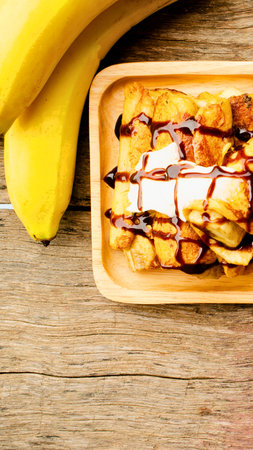 A close-up view of a sweet banana dessert topped with chocolate sauce, served on a wooden tray next to fresh bananas.の写真素材